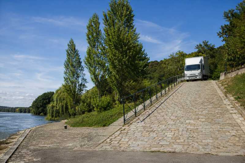 Rampe pavée descendant vers un quai bas en bord de Seine, camion de déménagement arrêté en haut de la pente