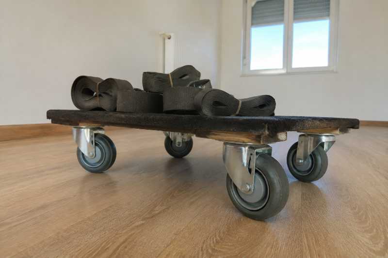 Chariot à piano bas à quatre roues pivotantes, sangles enroulées dessus, vide sur un parquet clair