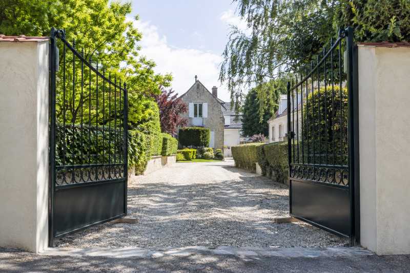 Portail à deux vantaux ouvert et maintenu par des cales de bois, allée de gravier dégagée derrière