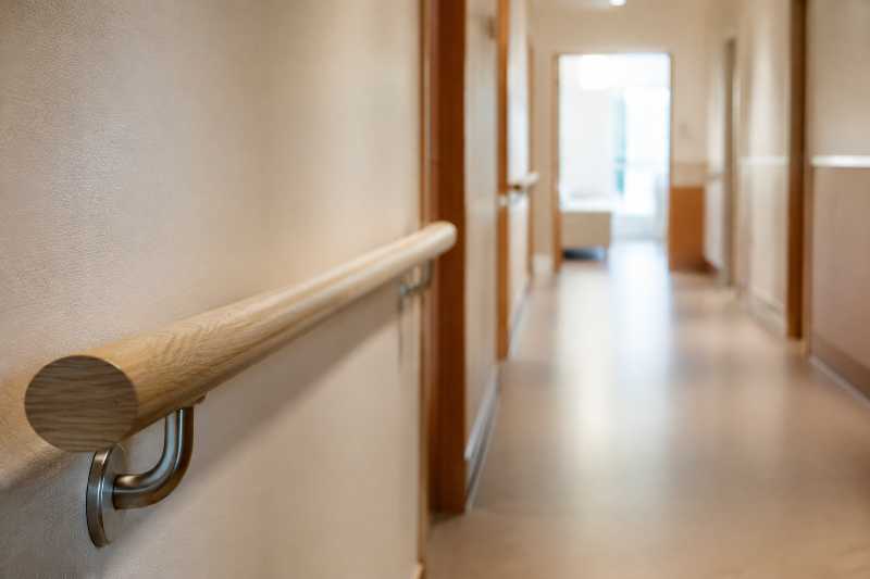 Main courante murale en bois clair le long d'un couloir d'établissement, porte de chambre ouverte au fond